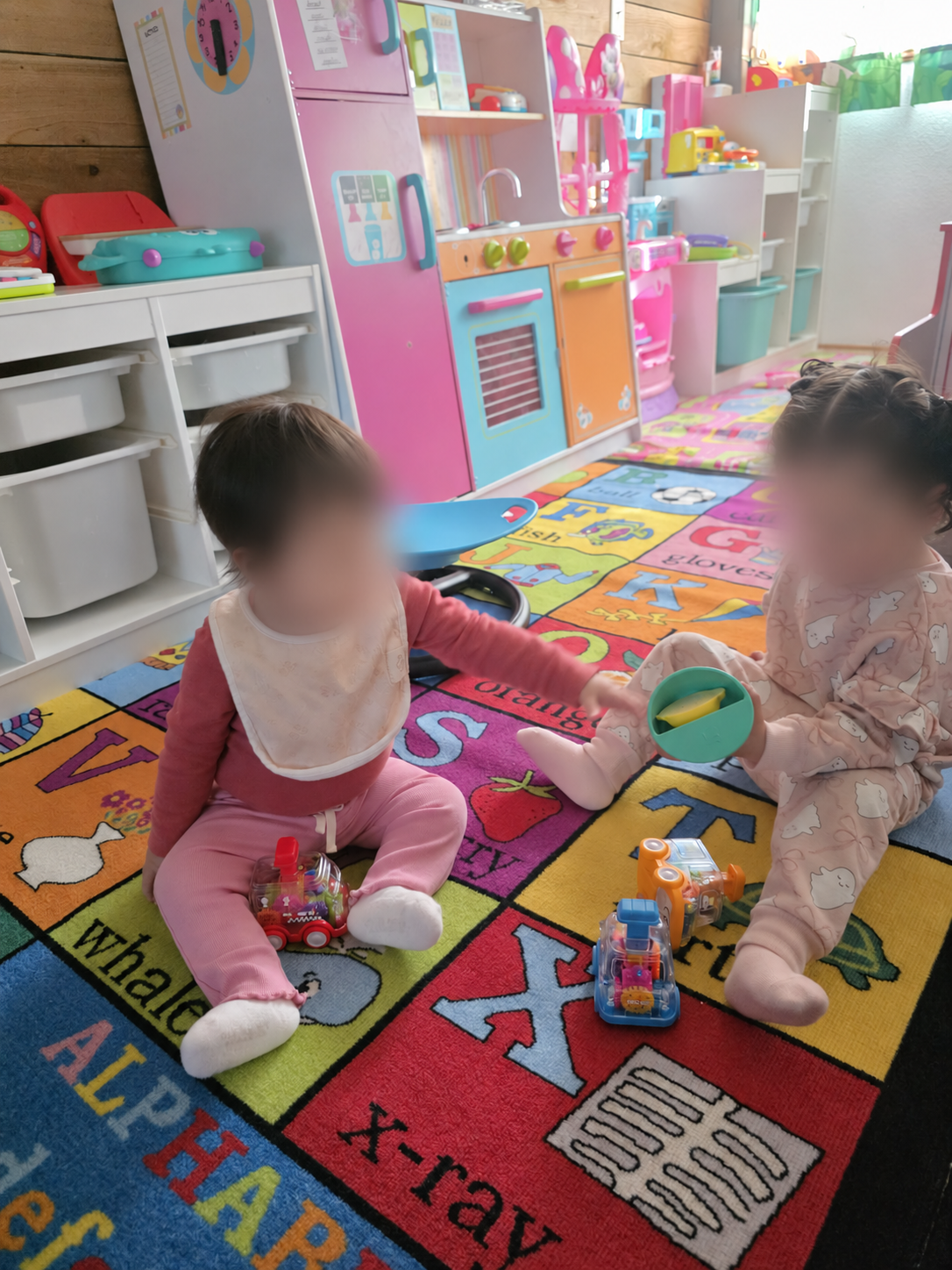 Little ones exploring toys on the playroom carpet, with faces blurred for privacy.